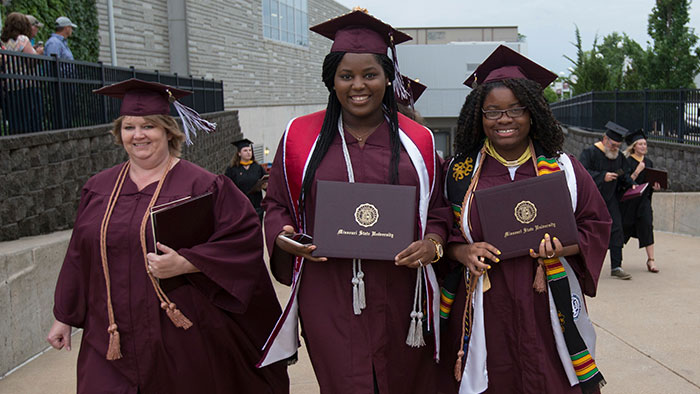 Three Missouri State graduates walking in their cap and gowns with their diplomas. 
